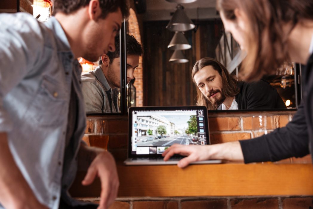 Cropped image of friends sitting in bar near the mirror and looking at laptop. Back view